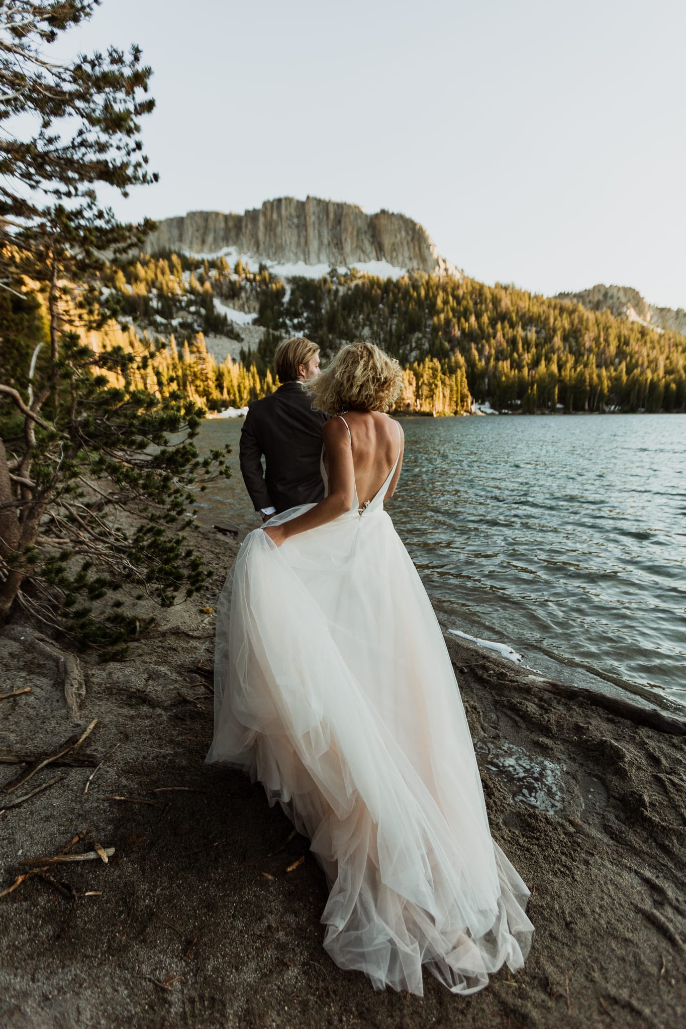 A bride and groom walk closely to the edge of McLeod in Mammoth Lakes. They are facing the tall Mammoth Crest ridge with the sun setting on them to their right.