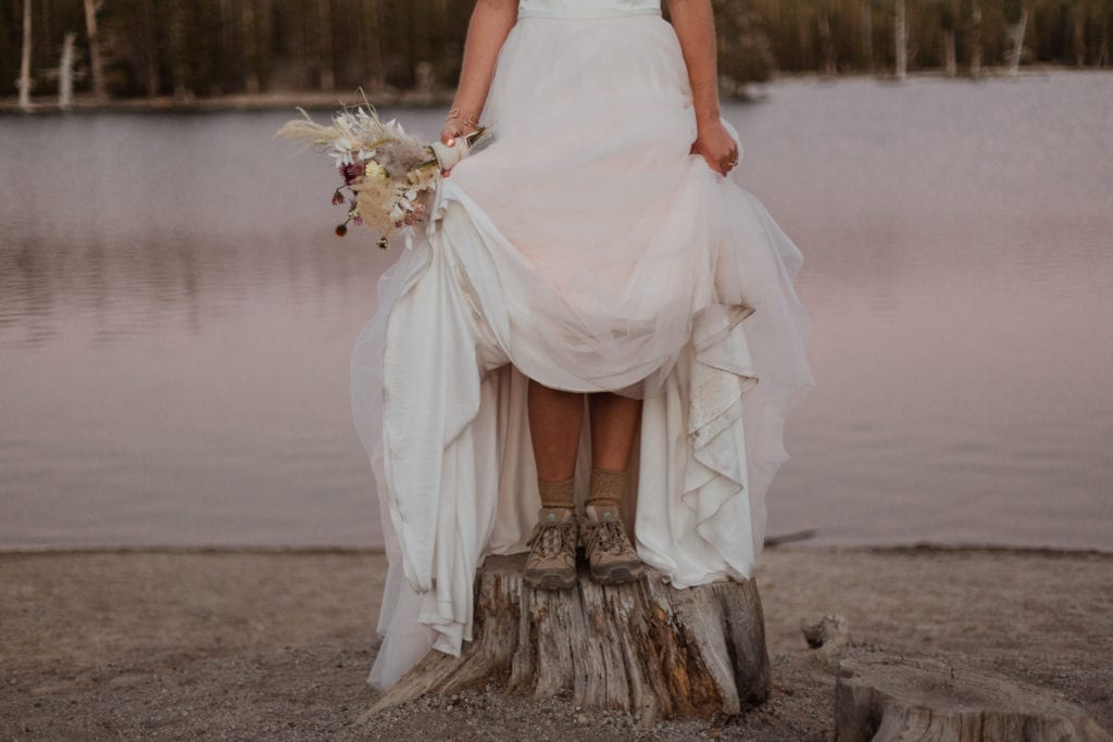 Haylee stands on a stump holding her wedding dress up so you can see her hiking shoes at Horseshoe Lake.