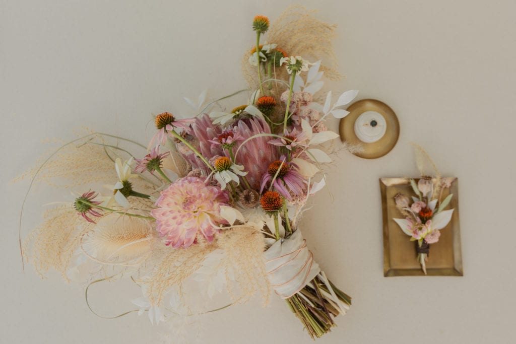 A bouquet of pink, white and orange flowers sit next to a wedding ring and boutonniere.